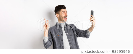 Profile of cheerful young guy taking selfie on smartphone, showing v-sign while make photo on mobile app, standing against white background 98395946