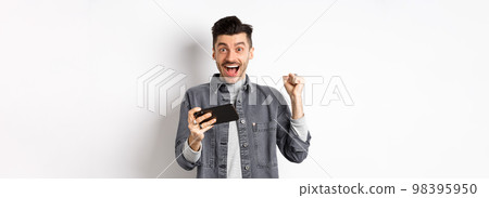 Excited man making money on mobile phone and rejoicing, raising hand up and scream from happiness and joy, standing with smartphone on white background Excited man making money on mobile phone and rejoicing, raising hand up and scream from happiness and joy, standing with smartphone on white background 98395950