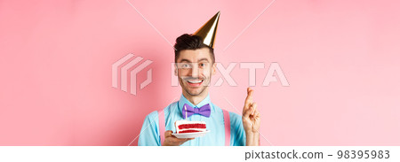 Holidays and celebration concept. Happy young man enjoying birthday party, wearing cone hat and cross fingers, making wish on bday cake with candle, pink background 98395983