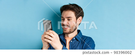 Close-up of handsome caucasian man writing message, reading mobile screen and smiling, standing on blue background Close-up of handsome caucasian man writing message, reading mobile screen and smiling, standing on blue background 98396002