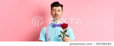 Cute and funny man waiting for kiss from lover on Valentines day, holding beautiful red rose for girlfriend, standing over pink background 98396080