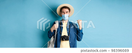 Covid-19 and summer holidays concept. Happy young man travelling abroad in medical mask, saying yes and raising fist up joyfully, cheerful about vacation, blue background Covid-19 and summer holidays concept. Happy young man travelling abroad in medical mask, saying yes and raising fist up joyfully, cheerful about vacation, blue background 98396089