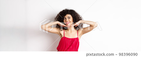 Beautiful lady with curly hair, wearing red dress, showing hear face with makeup and smiling, feeling carefree on white background Beautiful lady with curly hair, wearing red dress, showing hear face with makeup and smiling, feeling carefree on white background 98396090