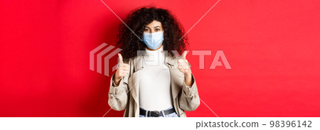 Covid-19, social distancing and quarantine concept. Cheerful stylish woman wearing trench coat and medical mask for going outside during pandemic, showing thumb up 98396142