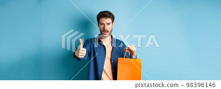Cool guy showing thumb up and holding orange shopping bag, standing on blue background 98396146