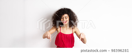 Cheerful elegant woman in red dress and makeup, looking and pointing down with pleased smile, showing advertisement, standing over white background Cheerful elegant woman in red dress and makeup, looking and pointing down with pleased smile, showing advertisement, standing over white background 98396281