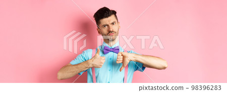 Skeptical young man in bow-tie grimacing, showing thumbs up and down, standing indecisive and judging something, standing over pink background Skeptical young man in bow-tie grimacing, showing thumbs up and down, standing indecisive and judging something, standing over pink background 98396283