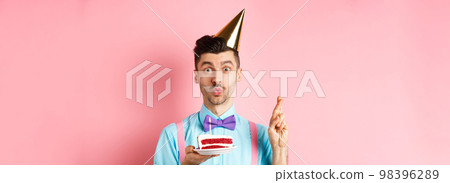 Holidays and celebration concept. Cute and funny man making birthday wish with crossed fingers, holding b-day cake and looking dreamy at camera, pink background Holidays and celebration concept. Cute and funny man making birthday wish with crossed fingers, holding b-day cake and looking dreamy at camera, pink background 98396289