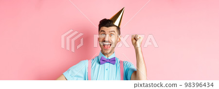 Holidays and celebration concept. Happy birthday boy with moustache and bow-tie celebrating event in party cone hat and festive clothes, raising hand up and shout yes Holidays and celebration concept. Happy birthday boy with moustache and bow-tie celebrating event in party cone hat and festive clothes, raising hand up and shout yes 98396434