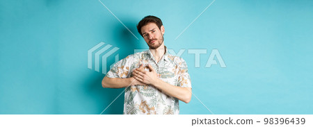 Passionate and romantic guy in hawaiian shirt, holding hands on heart and looking with sympathy, being in love, standing on blue background 98396439
