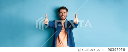 Handsome young man smiling and showing thumbs up in approval, say yes with happy face, standing on blue background 98397056