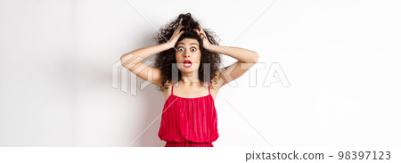 Shocked young woman holding hands on head and panicking, staring frustrated at camera, wearing red dress and makeup, white background 98397123