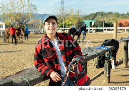 Portrait of positive young female stable worker holding horse halter or harness near wooden fence on ranch Portrait of positive young female stable worker holding horse halter or harness near wooden fence on ranch 98397179