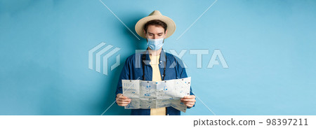 Covid-19, pandemic and travel concept. Young man in medical mask reading tourist map, searching for route to sightseeings, standing on blue background 98397211
