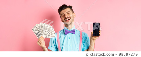 E-commerce and shopping concept. Rich and happy guy laughing, showing money in cash and empty smartphone screen, standing on pink background E-commerce and shopping concept. Rich and happy guy laughing, showing money in cash and empty smartphone screen, standing on pink background 98397279