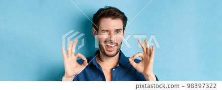 Close up of handsome confident guy winking, showing okay gestures, guarantee quality, recommending something good, standing on blue background Close up of handsome confident guy winking, showing okay gestures, guarantee quality, recommending something good, standing on blue background 98397322