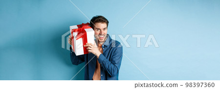 Valentines day. Happy young man got present on special holiday, trying guess what inside gift box and smiling, standing over blue background Valentines day. Happy young man got present on special holiday, trying guess what inside gift box and smiling, standing over blue background 98397360