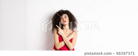 Indecisive silly woman in red dress, cross fingers and pointing sideways, looking up pensive, making choice, standing over white background 98397371
