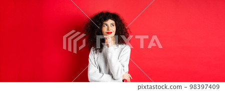 Pensive stylish woman with curly hair, red lips, looking thoughtful at upper left corner logo and smiling, standing against studio background Pensive stylish woman with curly hair, red lips, looking thoughtful at upper left corner logo and smiling, standing against studio background 98397409