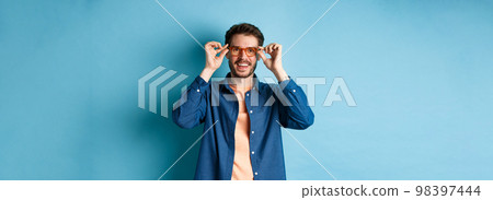 Happy caucasian man trying new glasses and smiling, standing on blue background 98397444