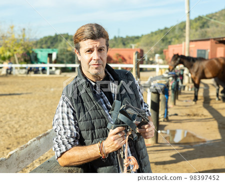 Middle-aged man instructor holding horse halter or reins during working day at horse club on sunny day of spring Middle-aged man instructor holding horse halter or reins during working day at horse club on sunny day of spring 98397452