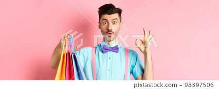 Funny guy in bow-tie showing OK sign and shopping bags, praising good deal in shops, standing over pink background Funny guy in bow-tie showing OK sign and shopping bags, praising good deal in shops, standing over pink background 98397506