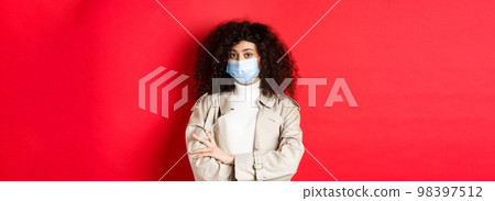 Covid-19, social distancing and quarantine concept. Portrait of skeptical girl in face mask and trench coat, cross arms on chest and staring unamused at camera, red background 98397512