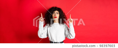 Concerned young woman with curly hair, frowning and looking doubtful, pointing up with hesitant or worried face, standing on red background Concerned young woman with curly hair, frowning and looking doubtful, pointing up with hesitant or worried face, standing on red background 98397603