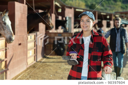 Portrait of young woman with brushes in her hands for cleaning and caring for horses at horse farm Portrait of young woman with brushes in her hands for cleaning and caring for horses at horse farm 98397604