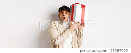 Holidays concept. Funny young man in glasses shaking gift box, listening what inside present and gasping amazed, standing on white background 98397605
