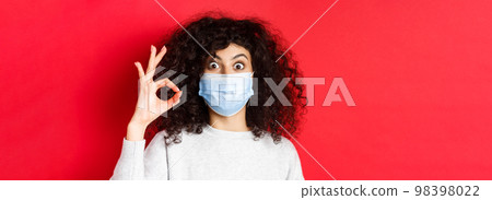 Health, pandemic and covid concept. Excited woman show yes okay sign, wearing medical mask and looking with approval, standing against red background 98398022