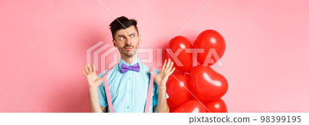 Valentines day concept. Pensive young man in bow-tie, raising hands up and looking left while thinking, making decision, standing on romantic pink background and heart balloons 98399195