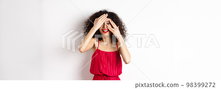 Surprise. Excited pretty girl with curly hair and red lips, peek through fingers and smiling amazed, checking out promo, standing on white background 98399272