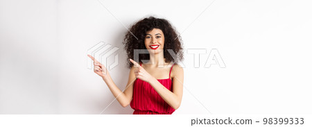 Cheerful female model in fashionable red dress, smiling and pointing fingers right at logo, standing over white background 98399333