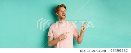 Dreamy handsome guy with red hair, smiling and looking at upper left corner with awe, admire something, standing over mint background 98399382