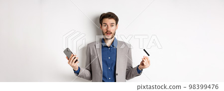 Online shopping. Confused man in suit spread hands sideways, holding plastic credit card with mobile phone and shrugging cueless, standing on white background 98399476