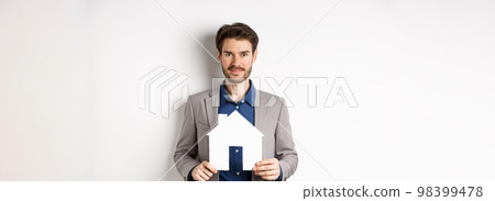 Real estate and insurance concept. Salesman in grey suit showing paper house cutout, selling property, smiling friendly at camera, white background Real estate and insurance concept. Salesman in grey suit showing paper house cutout, selling property, smiling friendly at camera, white background 98399478
