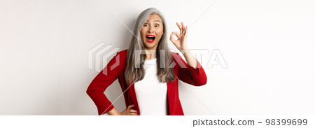 Happy asian mature woman in red blazer showing okay sign, smiling amazed, praising something awesome, standing over white background Happy asian mature woman in red blazer showing okay sign, smiling amazed, praising something awesome, standing over white background 98399699