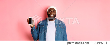 Handsome hipster guy in beanie and denim shirt smiling, showing mobile phone screen with happy face, introduce application, standing over pink background Handsome hipster guy in beanie and denim shirt smiling, showing mobile phone screen with happy face, introduce application, standing over pink background 98399701