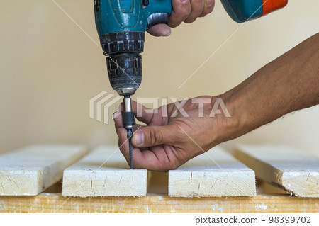 Hand of a worker screws a screw in a wooden board with a cordless screwdriver. Man carpenter at handmade work Hand of a worker screws a screw in a wooden board with a cordless screwdriver. Man carpenter at handmade work 98399702