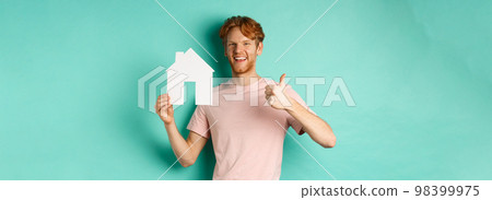 Real estate concept. Young man with red hair, wearing t-shirt, showing paper house cutout and thumb up, recommend agency, standing over turquoise background 98399975