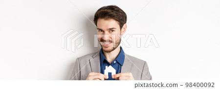 Real estate and insurance concept. Smiling male realtor showing small paper house cutout and looking at camera, selling property, white background Real estate and insurance concept. Smiling male realtor showing small paper house cutout and looking at camera, selling property, white background 98400092