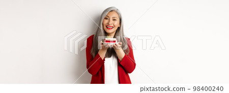 Beautiful asian grandmother holding piece of cake and smiling, standing in red blazer over white background Beautiful asian grandmother holding piece of cake and smiling, standing in red blazer over white background 98400240