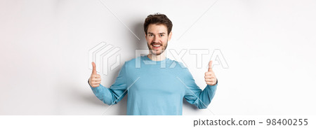 Cheerful young man showing thumbs up and smiling, recommend good product, praising choice, standing on white background 98400255