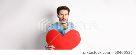 Romantic boyfriend sending air kiss at camera and holding big red Valentines day heart, standing over white background 98400525