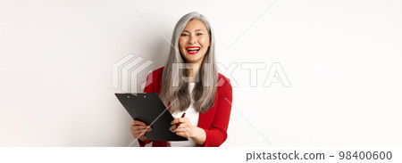 Happy asian office employee, woman in red blazer, working and laughing, holding clipboard with documents, standing over white background Happy asian office employee, woman in red blazer, working and laughing, holding clipboard with documents, standing over white background 98400600