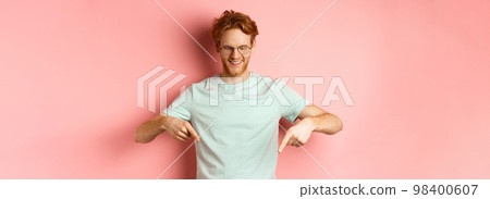Image of handsome caucasian man with red messy hair and glasses, pointing fingers and looking down at promo offer, smiling pleased, standing over pink background Image of handsome caucasian man with red messy hair and glasses, pointing fingers and looking down at promo offer, smiling pleased, standing over pink background 98400607