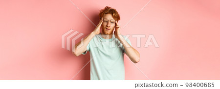 Portrait of redhead man in crooked glasses touching head and feeling dizzy or nauseous, having hangover or headache, standing over pink background 98400685