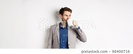 Handsome businessman in suit drinking coffee or tea from office mug, looking aside at logo, standing against white background Handsome businessman in suit drinking coffee or tea from office mug, looking aside at logo, standing against white background 98400716