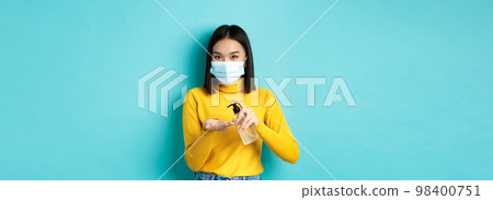 Covid-19, social distancing and pandemic concept. Smiling asian woman in medical mask using preventive measures from coronavirus, using hand sanitizer, standing over blue background Covid-19, social distancing and pandemic concept. Smiling asian woman in medical mask using preventive measures from coronavirus, using hand sanitizer, standing over blue background 98400751